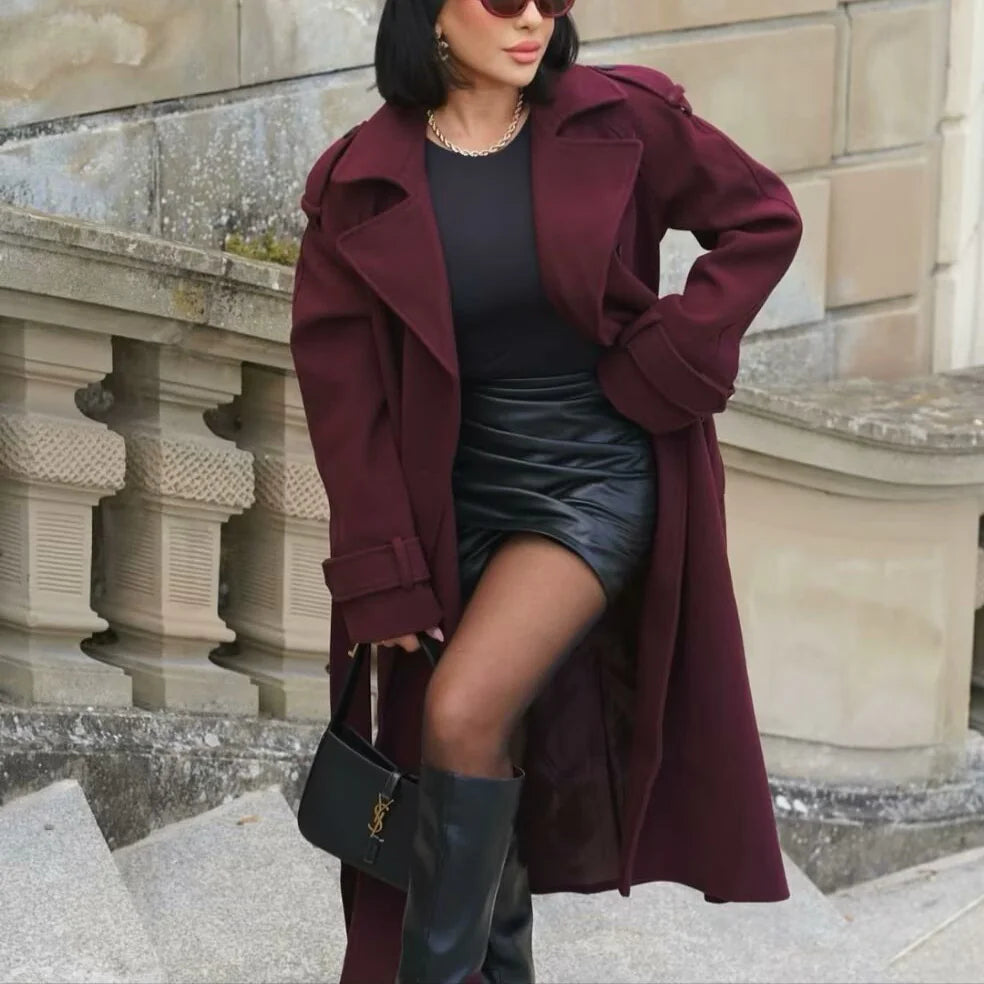 Woman wearing a burgundy coat, black top, and skirt with knee-high boots, standing against a stone wall.