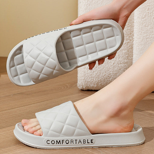 White quilted slide sandals with 'COMFORTABLE' text worn by a person on a wooden floor.